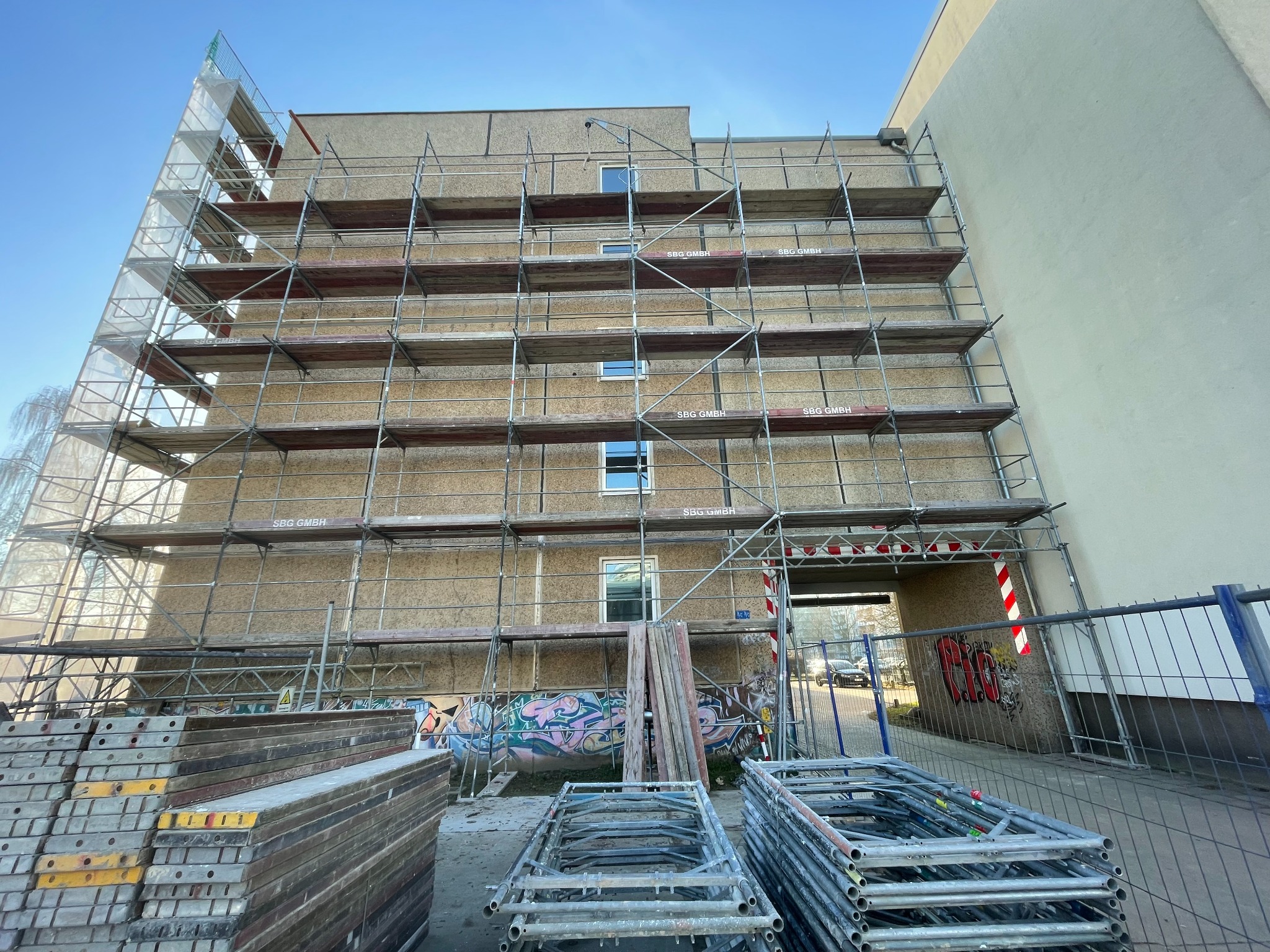 Siedmiopiętrowy budynek w trakcie renowacji z rusztowaniem, widoczne stosy elementów rusztowania na pierwszym planie, graffiti na ścianie budynku pod rusztowaniem.