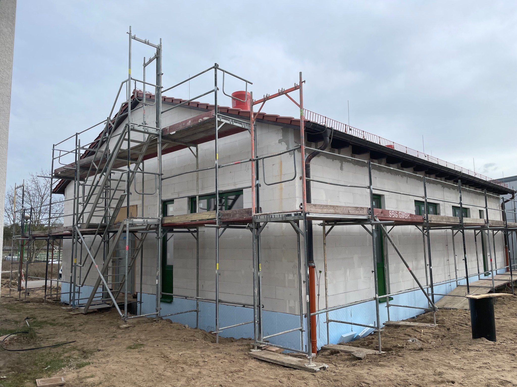 Budynek w trakcie budowy z białych bloczków, otoczony metalowym rusztowaniem, widoczne zielone ramy okienne i niebieska izolacja fundamentów.