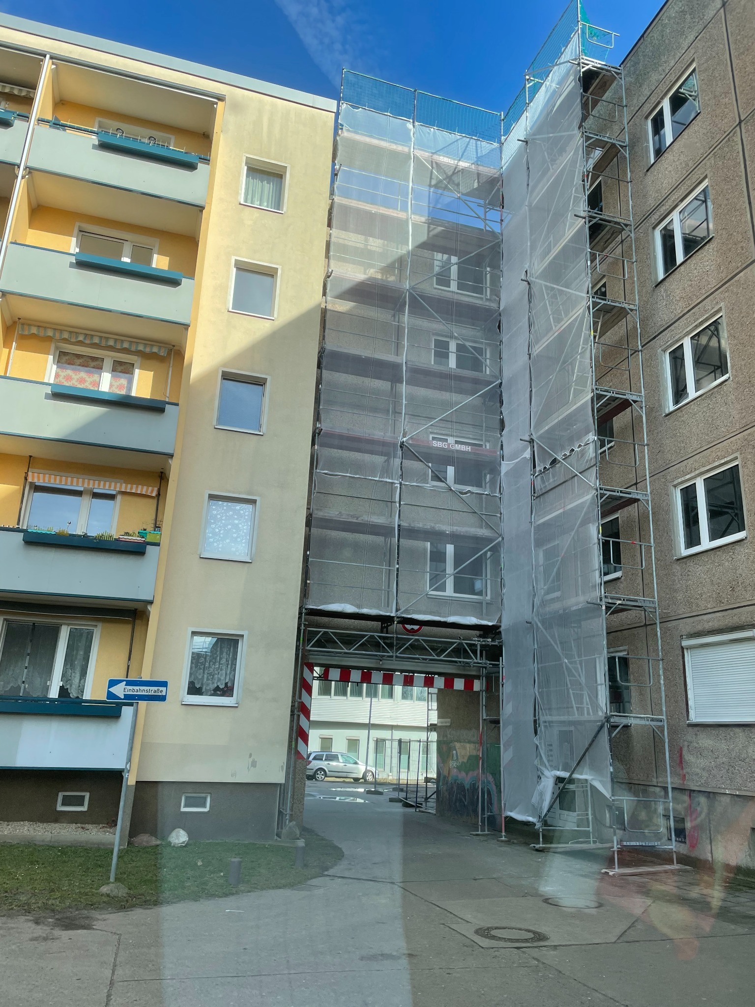 Dwa budynki mieszkalne połączone rusztowaniem z siatką ochronną, widoczne okna i balkony.