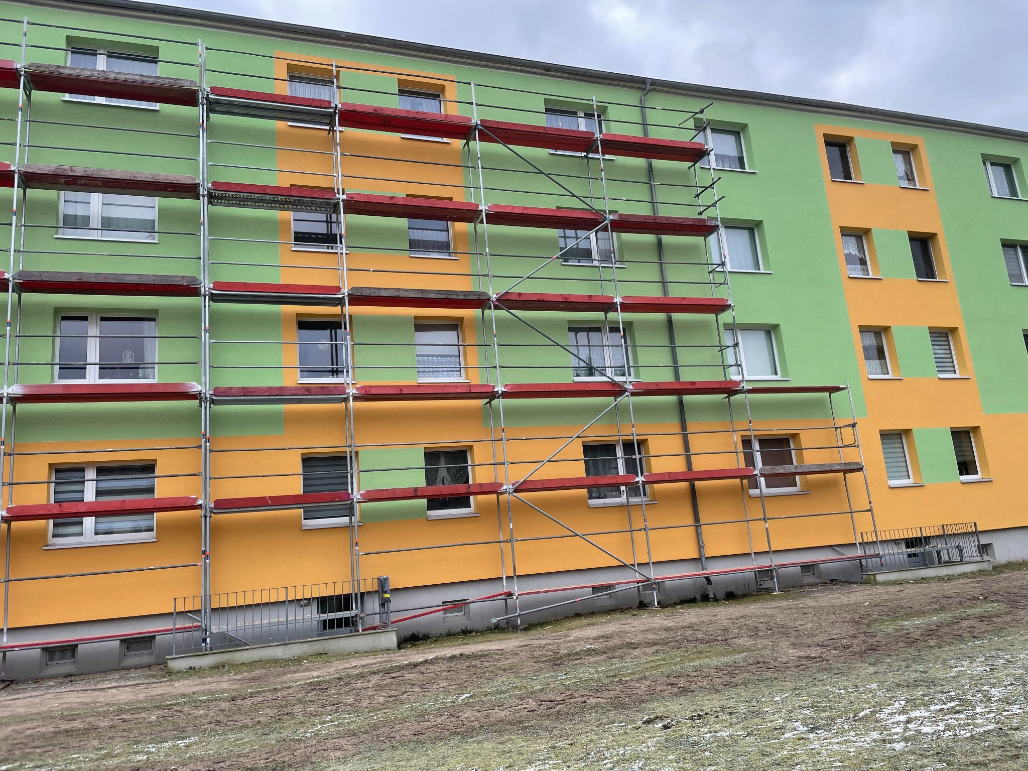 Wielopiętrowy budynek w trakcie malowania elewacji na zielono i żółto, z widocznym rusztowaniem na całej ścianie.