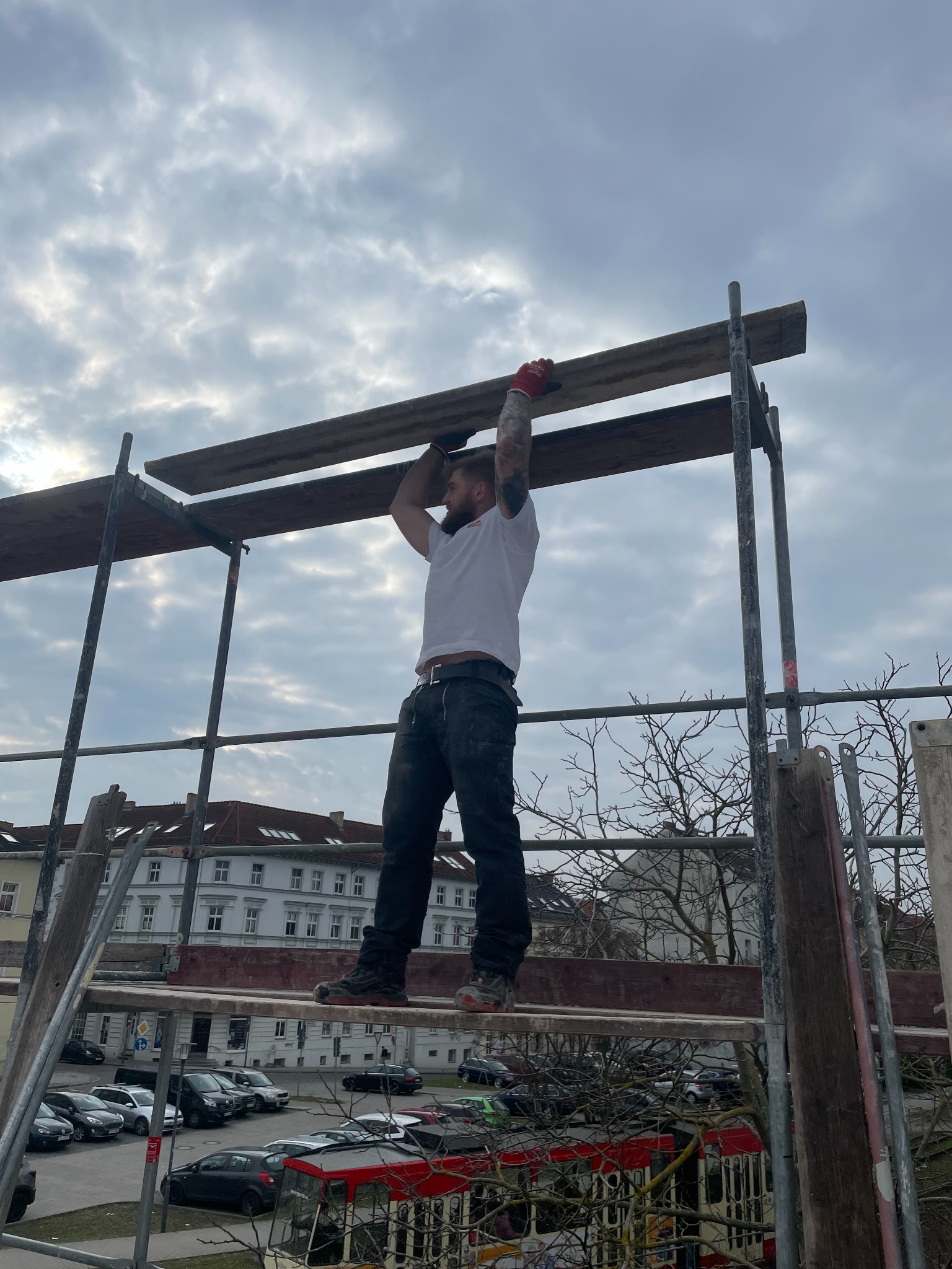 Mężczyzna z tatuażami na rusztowaniu układa deski, widok z dołu na tle pochmurnego nieba i zabudowań miejskich.
