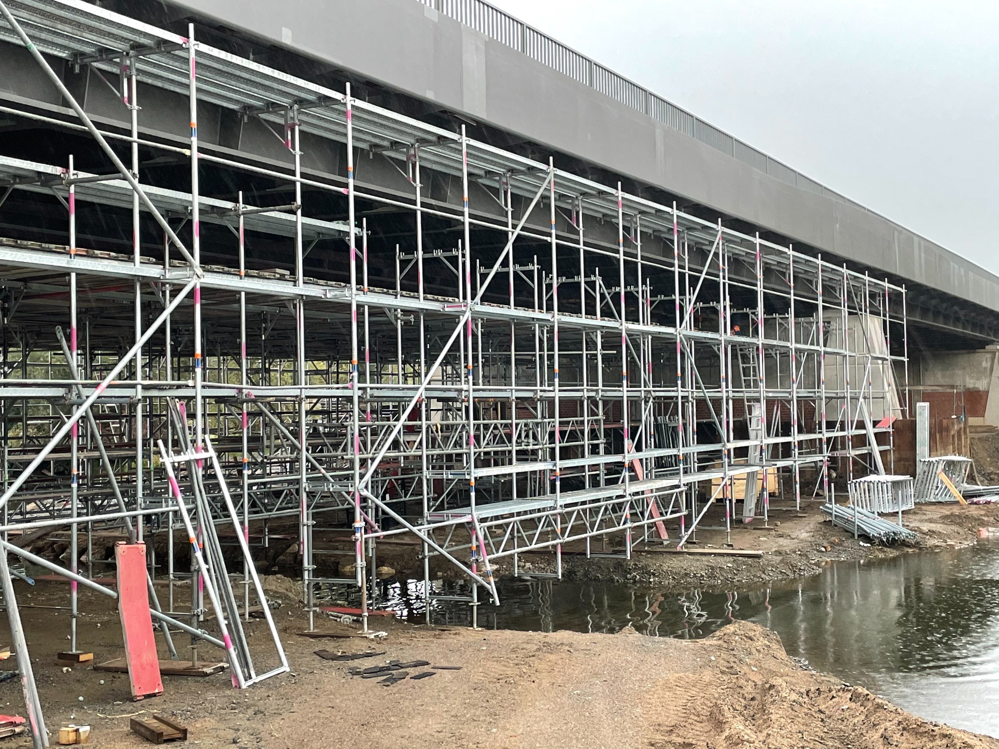 Rozbudowane rusztowanie z metalowych rur podtrzymujące most, częściowo zanurzone w wodzie, z widocznymi elementami konstrukcyjnymi i błotnistym brzegiem.