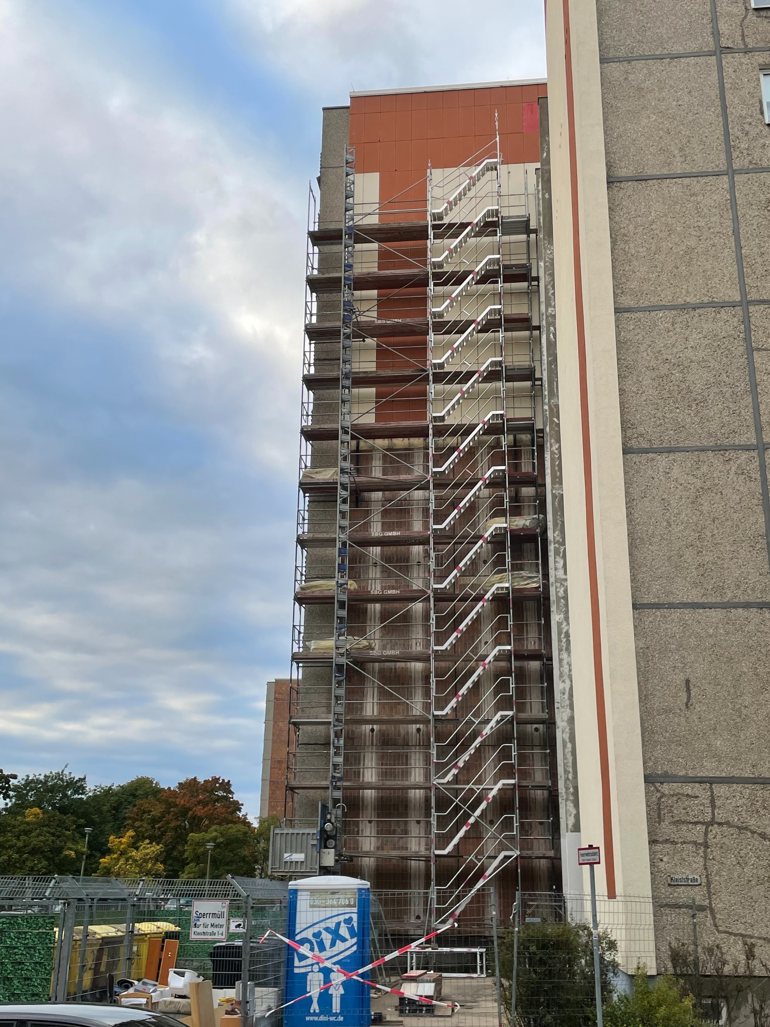 Wysoki budynek w trakcie renowacji z zamontowanym rusztowaniem na całej wysokości, widoczna przenośna toaleta na dole i fragment tablicy informacyjnej.