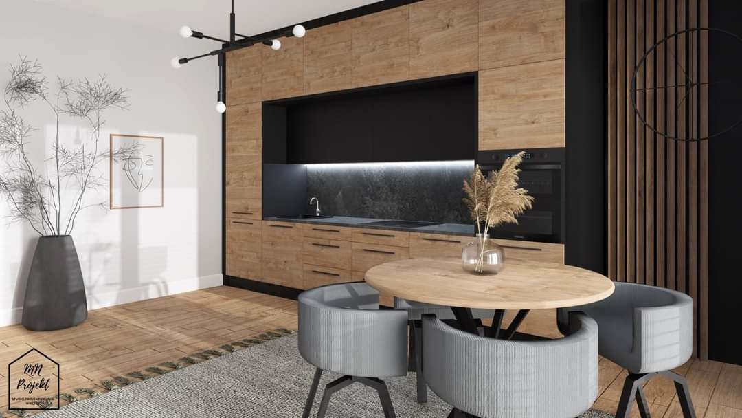 Wnętrze kuchni z drewnianymi szafkami, czarnymi akcentami i okrągłym stołem z szarymi krzesłami, oświetlone nowoczesną lampą wiszącą. Na stole wazon z suchymi trawami. Na ścianie minimalistyczny...