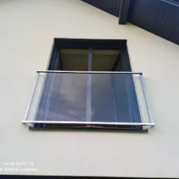 Zewnętrzny balkon z przezroczystą szklaną barierką i aluminiową ramą, widok z dołu na tle jasnej elewacji budynku.