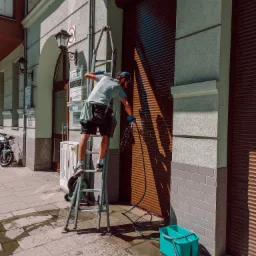 Mężczyzna na drabinie w niebieskich rękawicach, spryskuje roletę budynku wodą z węża, obok stoi turkusowe wiadro, a w tle widać zaparkowany motocykl.