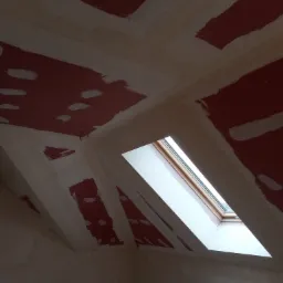 Skośny sufit poddasza w trakcie szpachlowania w Krakowie, widoczne fragmenty czerwonej farby i okno dachowe.