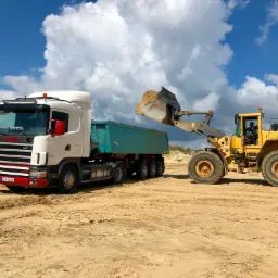 Żółta ładowarka Volvo nabiera urobek do turkusowej naczepy ciężarówki Scania z czerwoną tablicą rejestracyjną ZKA 38848 na piaszczystym placu budowy pod błękitnym niebem z białymi chmurami.