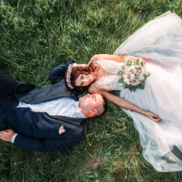 Para młoda w strojach ślubnych leży na trawie z widokiem z góry, panna młoda w koronie z kwiatów i z bukietem w ręku patrzy w obiektyw.