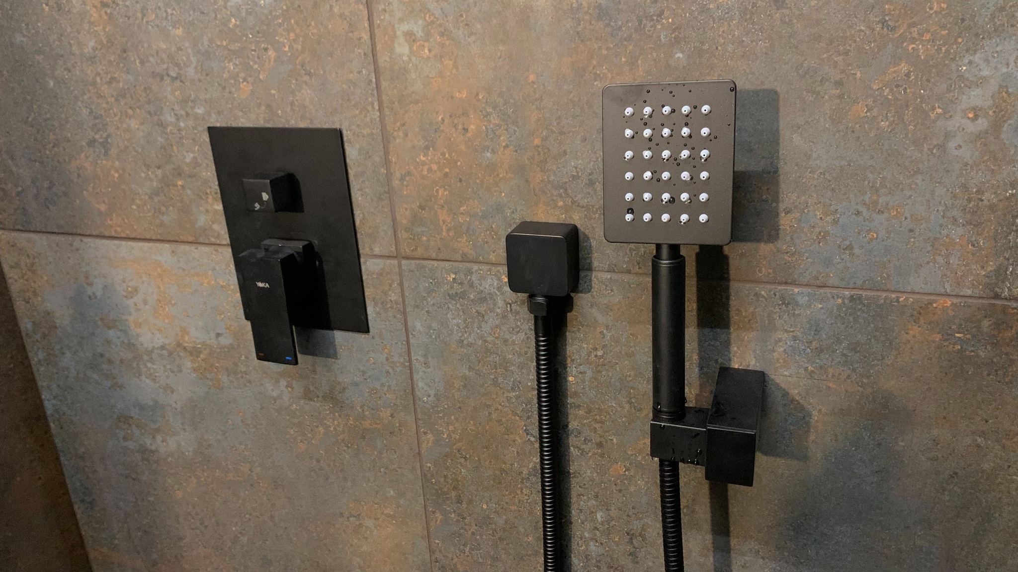 Matowa, czarna bateria prysznicowa z deszczownicą i wylewką zamontowana na ścianie z płytek imitujących rdzę.