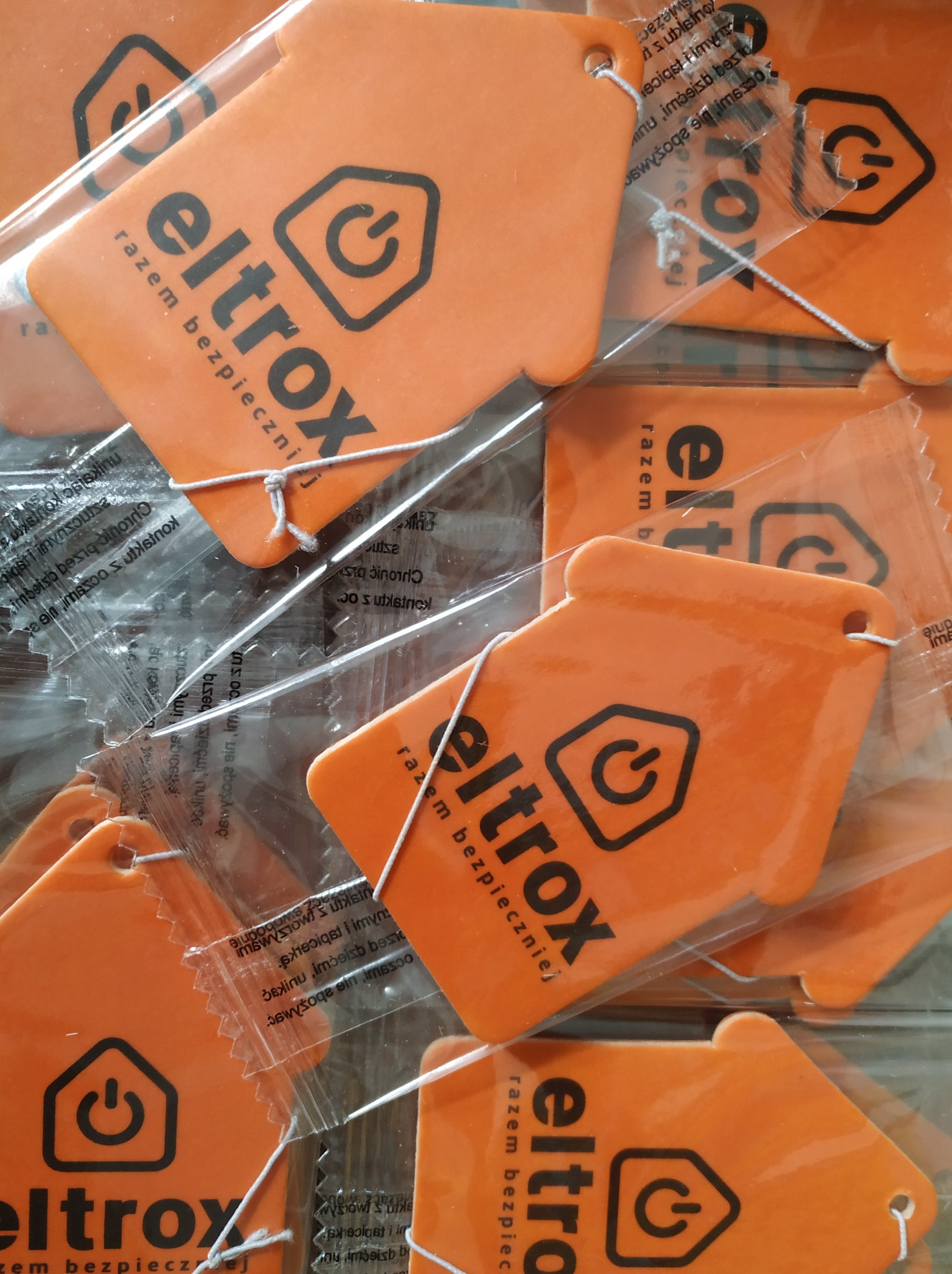 Pomarańczowe zawieszki zapachowe w kształcie domku z logo 'eltrox razem bezpieczniej' w przezroczystych opakowaniach, ułożone blisko siebie.