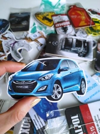 Niebieski Hyundai i30 jako naklejka trzymana w dłoni, otoczony innymi naklejkami z motywami motoryzacyjnymi i sportowymi na białym tle.