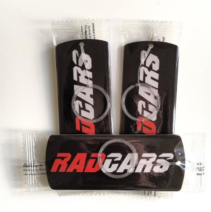 Trzy zapakowane odświeżacze powietrza do samochodu z logo 'RAD CARS' w kolorach czerwieni i bieli na czarnym tle, z plastikowymi zawieszkami.