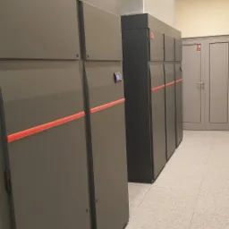 Rząd szarych szaf z poziomymi czerwonymi paskami w pomieszczeniu technicznym z widoczną instalacją wentylacyjną pod sufitem i podłogą z płytek.