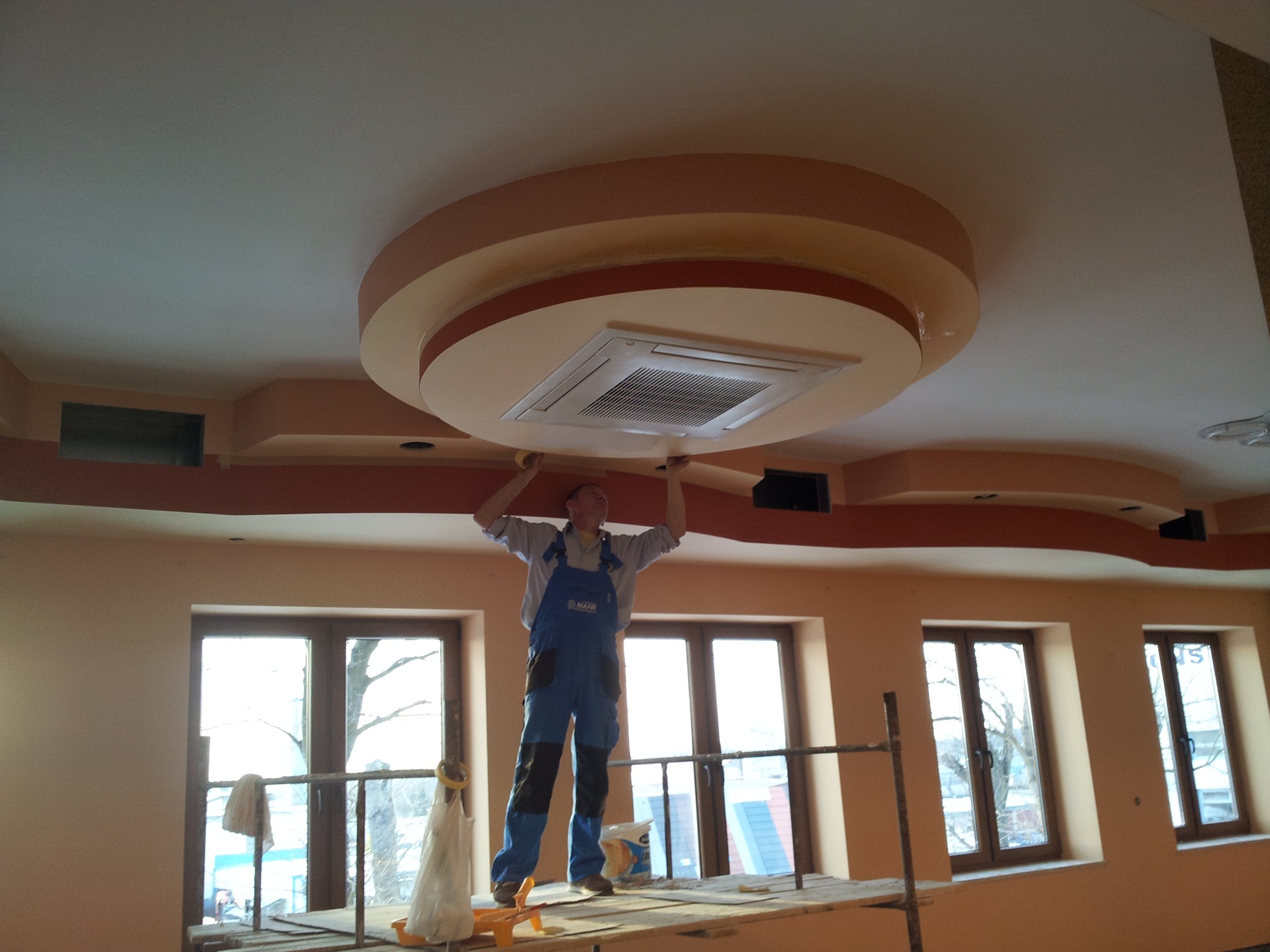 Instalator na rusztowaniu montuje klimatyzację kasetonową w ozdobnym suficie podwieszanym. Widoczne okna i pomalowane ściany w pomieszczeniu.