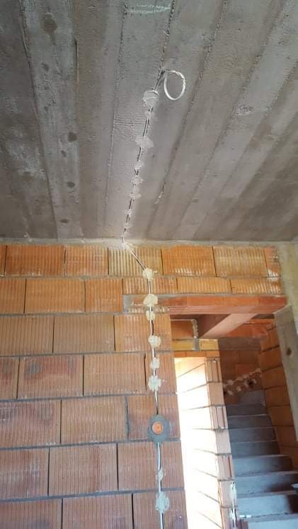 Wewnętrzna instalacja elektryczna w surowym stanie: przewód przymocowany do sufitu i ściany z cegły za pomocą uchwytów, widoczne puszki instalacyjne i schody w tle.