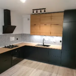 Nowoczesna kuchnia z czarnymi szafkami dolnymi i drewnianymi górnymi, oświetlenie punktowe, szara ściana nad blatem.