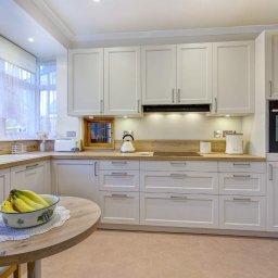 Teoria Wnętrza Aleksandra Koter - Jasna kuchnia z białymi szafkami, drewnianym blatem i okrągłym stołem z owocami w misce. Uwagę przyciąga małe okienko w ścianie nad blatem.