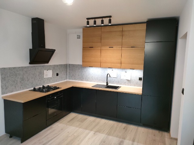 Nowoczesna kuchnia z czarnymi szafkami dolnymi i drewnianymi górnymi, oświetlenie punktowe, szara ściana nad blatem.