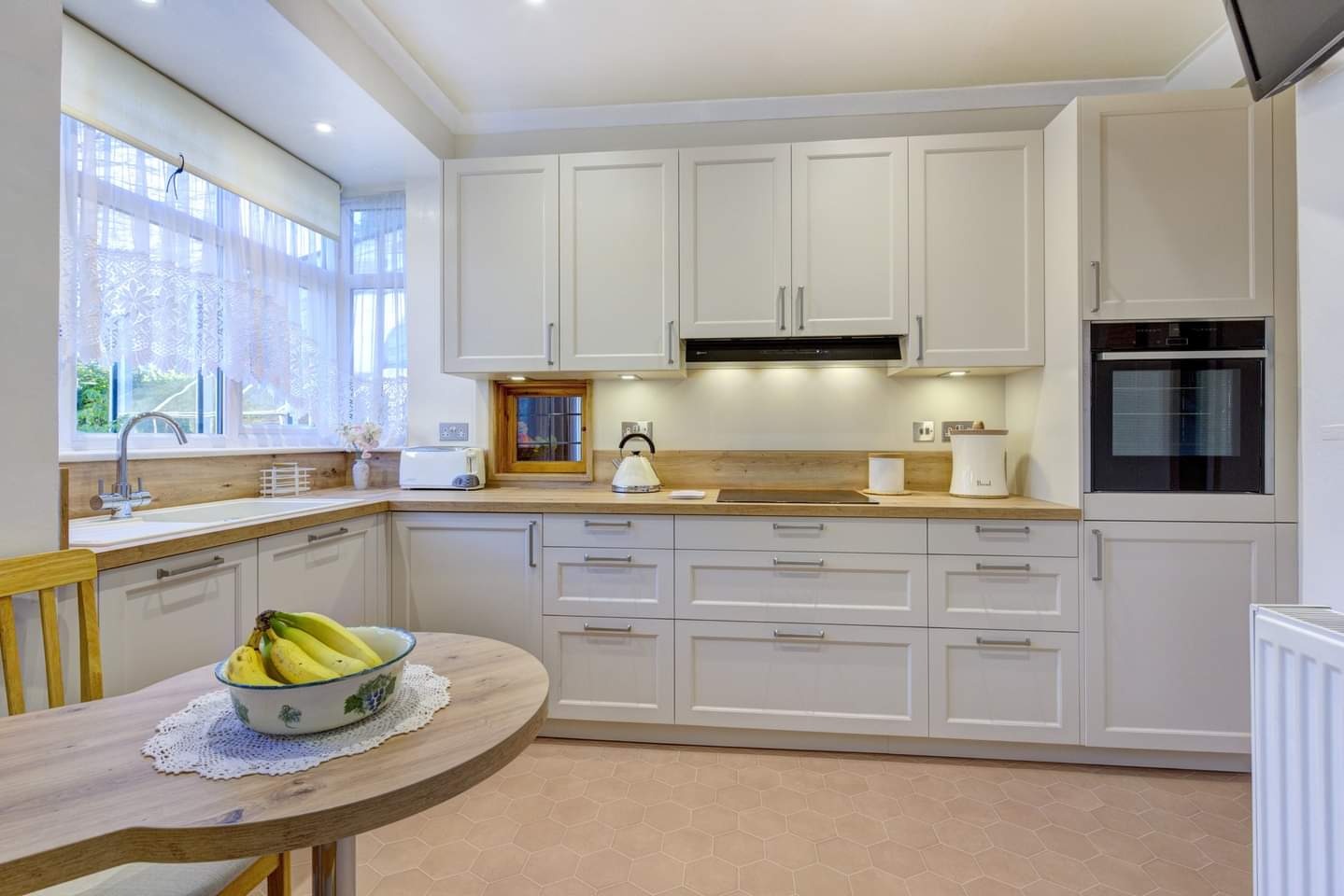 Jasna kuchnia z białymi szafkami, drewnianym blatem i okrągłym stołem z owocami w misce. Uwagę przyciąga małe okienko w ścianie nad blatem.