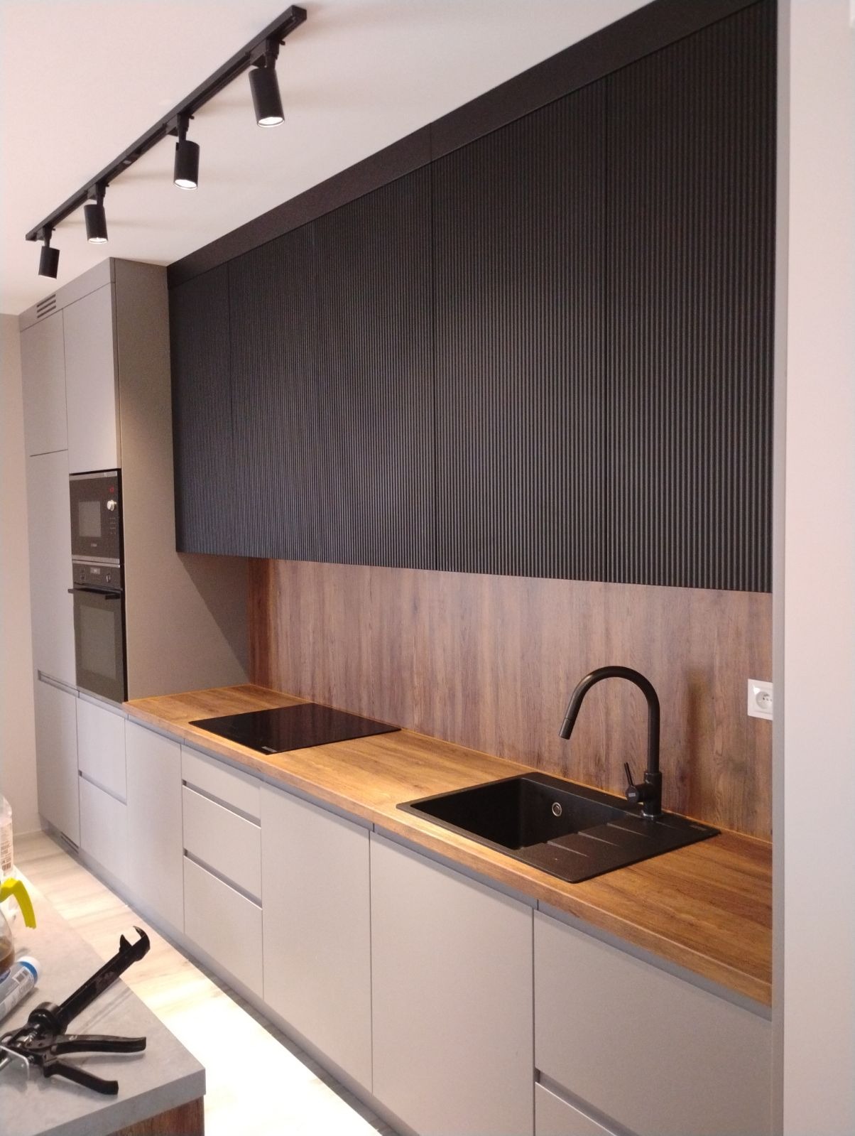 Szara zabudowa kuchenna z drewnianym blatem, czarnym zlewem i panelem ściennym imitującym drewno, oświetlona reflektorami na szynie.