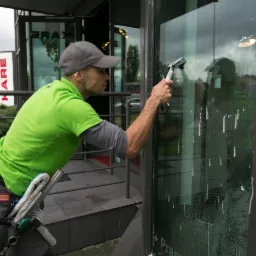 Pracownik w zielonej koszulce, z boku, czyści okno budynku w Krakowie za pomocą ściągaczki, widoczne ślady wody i piany na szkle, w tle fragment ulicy i zabudowań.