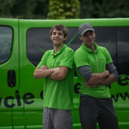 Dwaj uśmiechnięci pracownicy firmy X-Clean w zielonych koszulkach polo stoją na tle zielonego samochodu dostawczego z napisem 'Mycie elewacji' i adresem strony internetowej firmy.