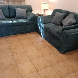 Komplet wypoczynkowy: sofa dwuosobowa i fotel w odcieniach zieleni, z poduszkami w paski, na tle jasnych płytek podłogowych i lampy.