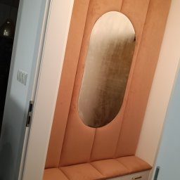 IM Meble Iwona Ziarkowska - Tapicerowany panel ścienny w odcieniach brzoskwini z owalnym lustrem i siedziskiem z szafkami w przedpokoju. Złote uchwyty dodają elegancji.