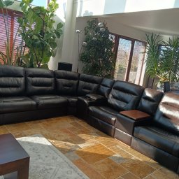 IM Meble Iwona Ziarkowska - Czarna, skórzana sofa narożna z funkcją relaks w jasnym salonie z dużymi oknami i roślinami. Widoczny stolik kawowy i dywan. Sofa po renowacji.