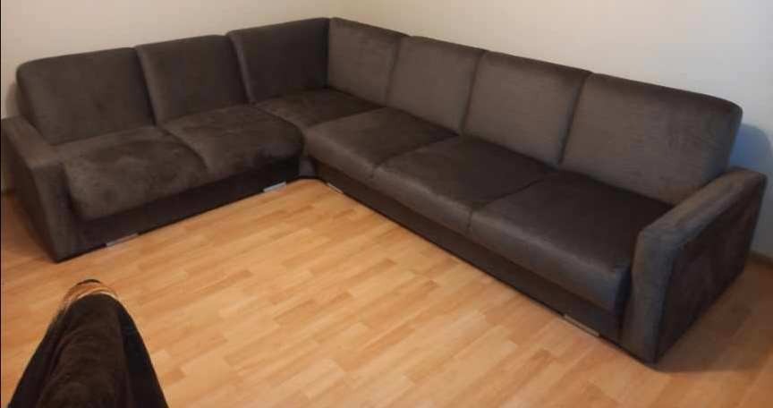 Ciemnobrązowy narożnik z materiałową tapicerką na jasnej drewnianej podłodze, widoczne ślady użytkowania na siedzisku.