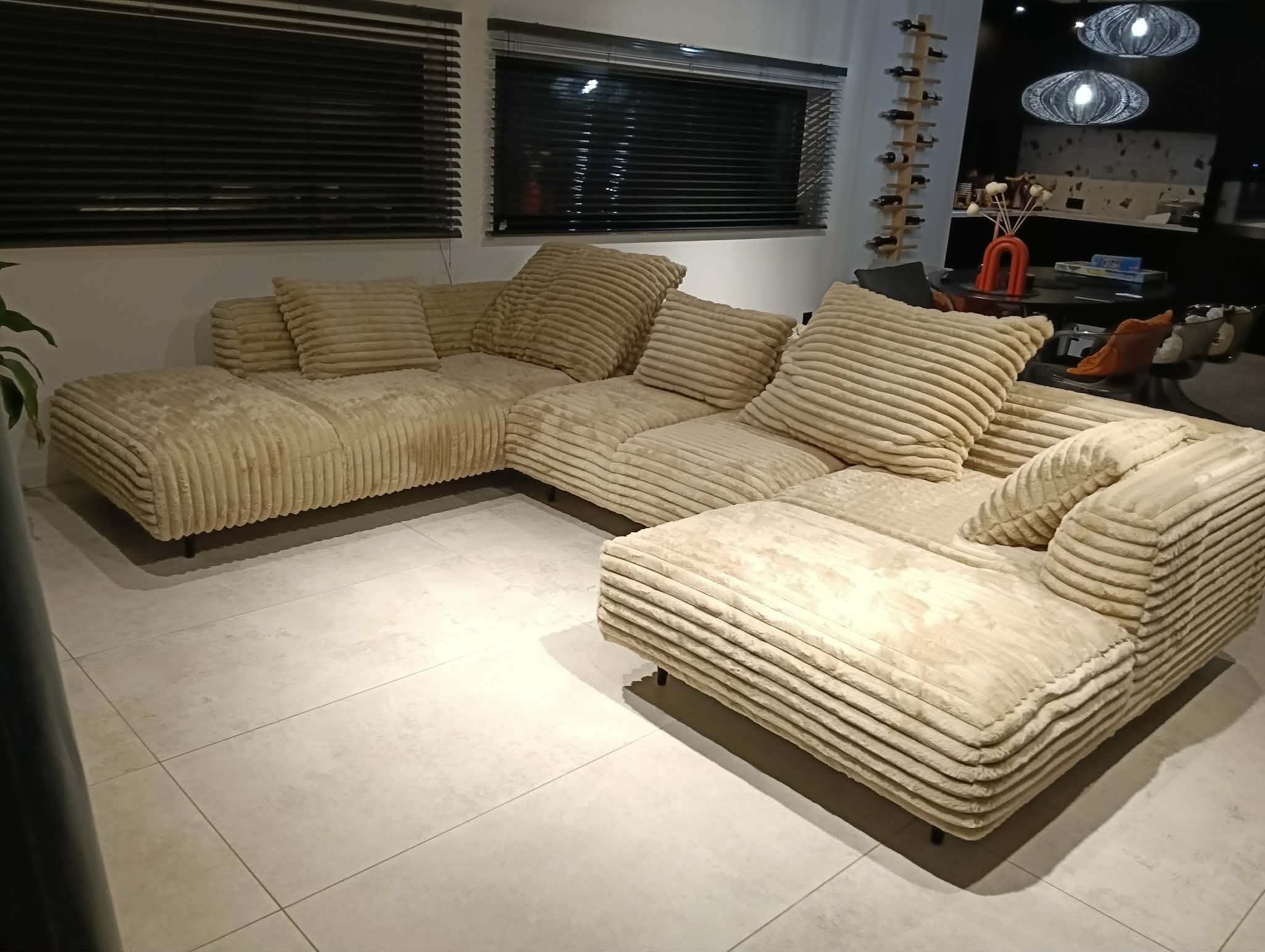 Beżowy, modułowy narożnik z tkaniny w prążki, z poduszkami, na tle okna z żaluzjami i elementami dekoracyjnymi wnętrza. Nowoczesny design.