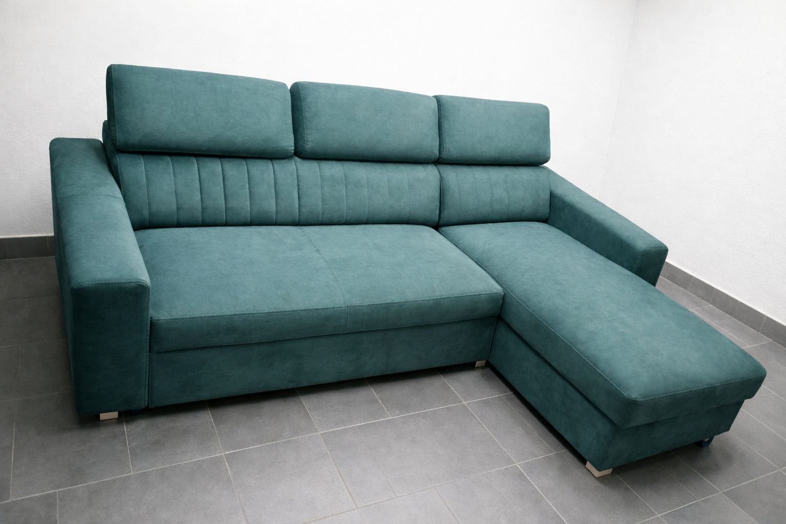 Turkusowa sofa narożna z regulowanymi zagłówkami, obita miękką tkaniną, na tle szarej podłogi i białej ściany. Widoczne subtelne nóżki.