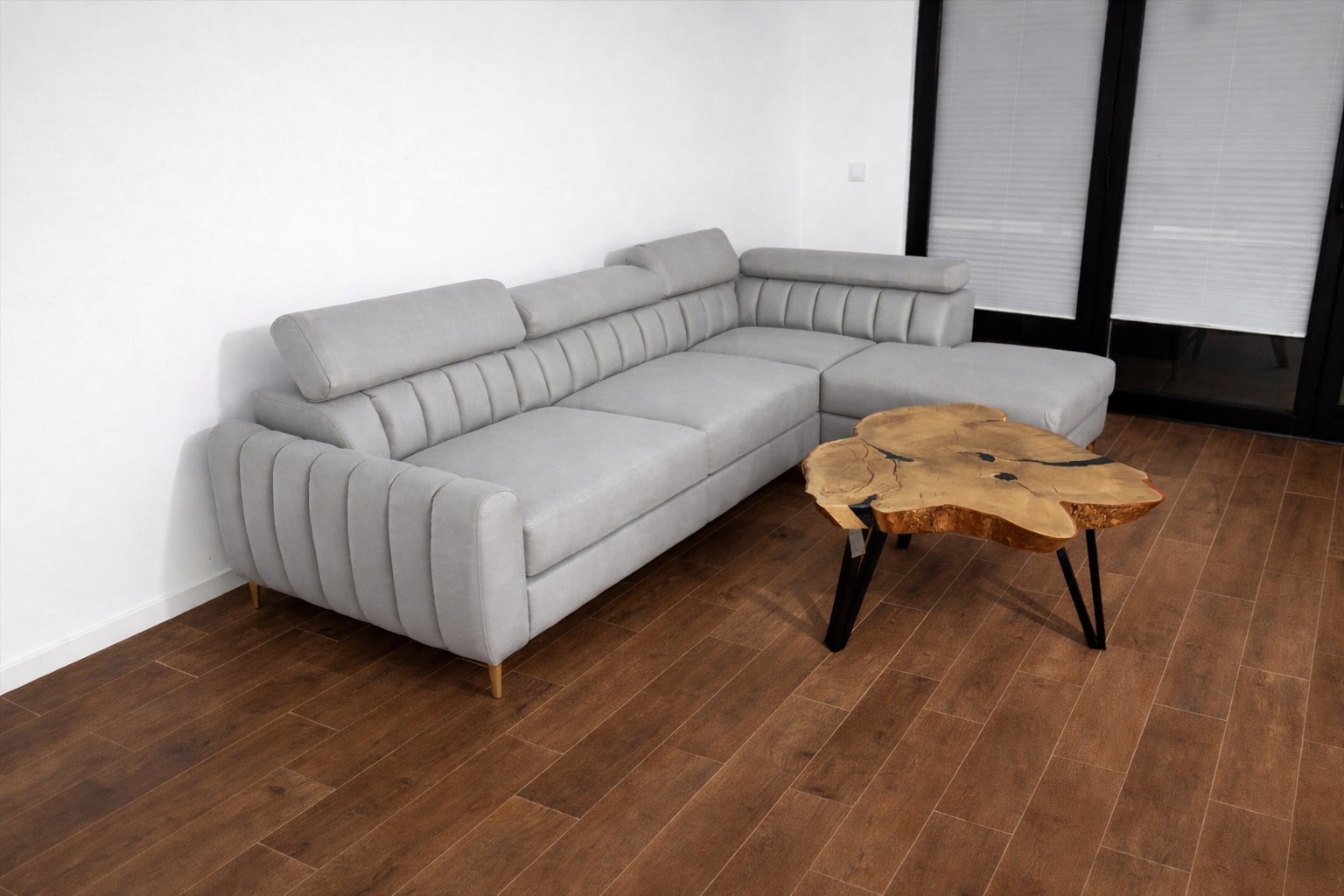 Szary narożnik z pikowanymi bokami i drewniany stolik kawowy na tle brązowej podłogi i okna z żaluzjami. Nowoczesny design mebli tapicerowanych.