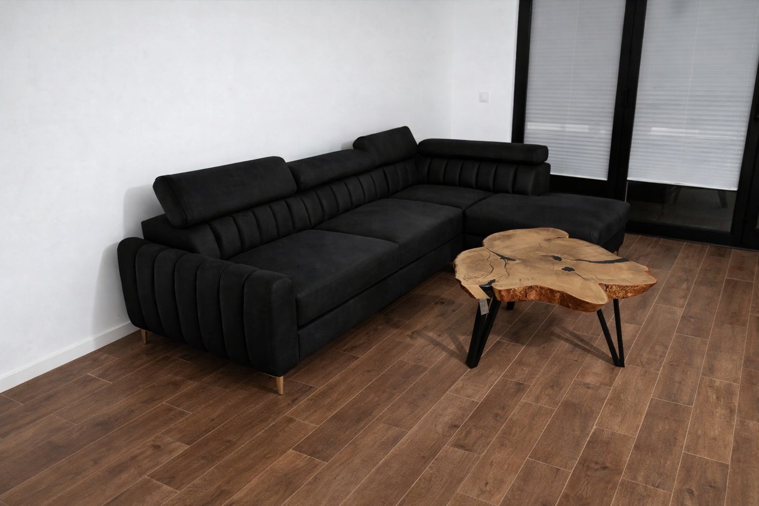 Czarny, welurowy narożnik z regulowanymi zagłówkami, na drewnianej podłodze, obok stolik z blatem z plastra drewna. Nowoczesny design, jasne wnętrze.