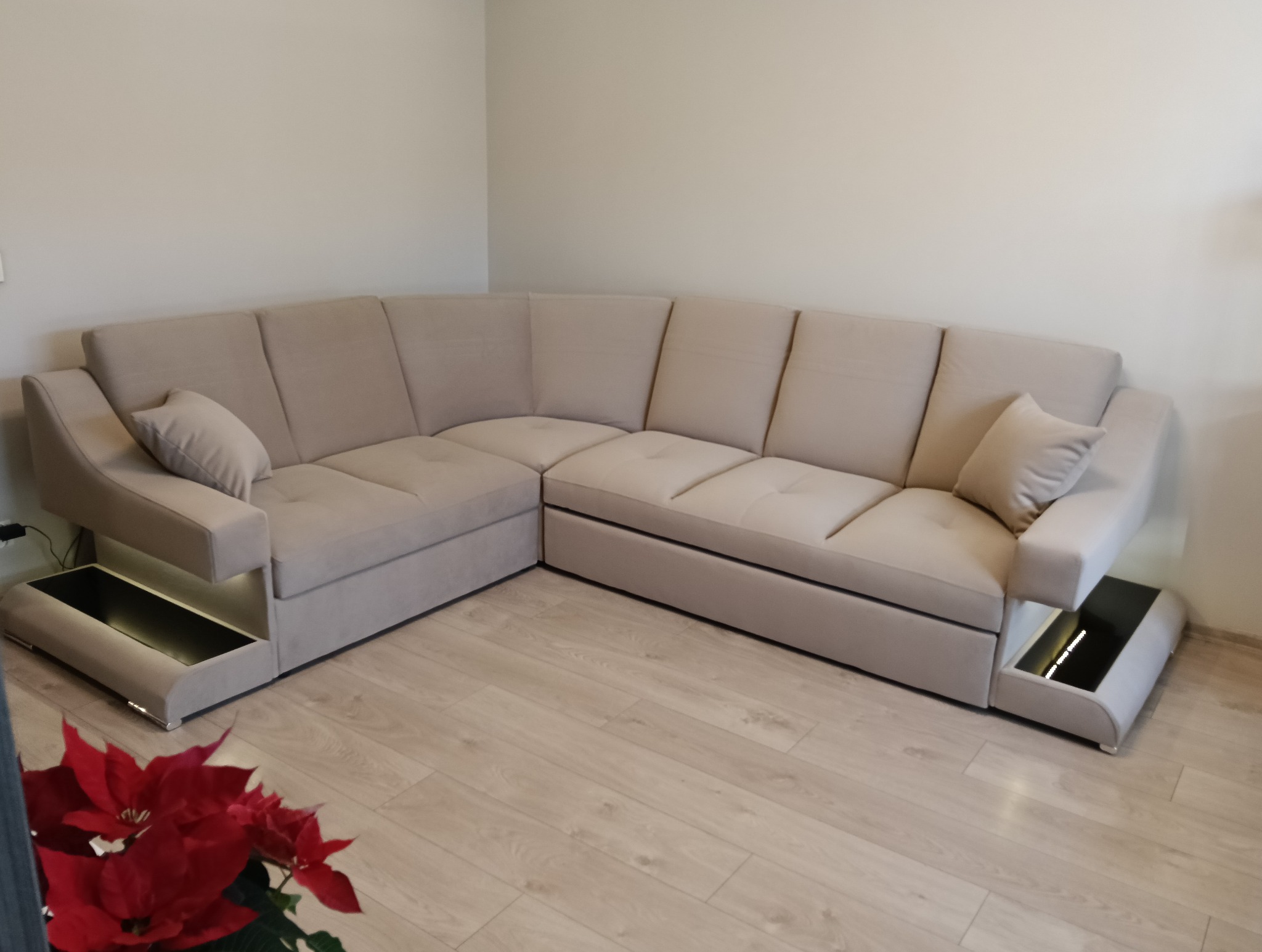 Beżowy narożnik z podświetlanymi półkami bocznymi, ustawiony na jasnej podłodze. Widoczny fragment czerwonej rośliny w lewym dolnym rogu. Minimalistyczny design.