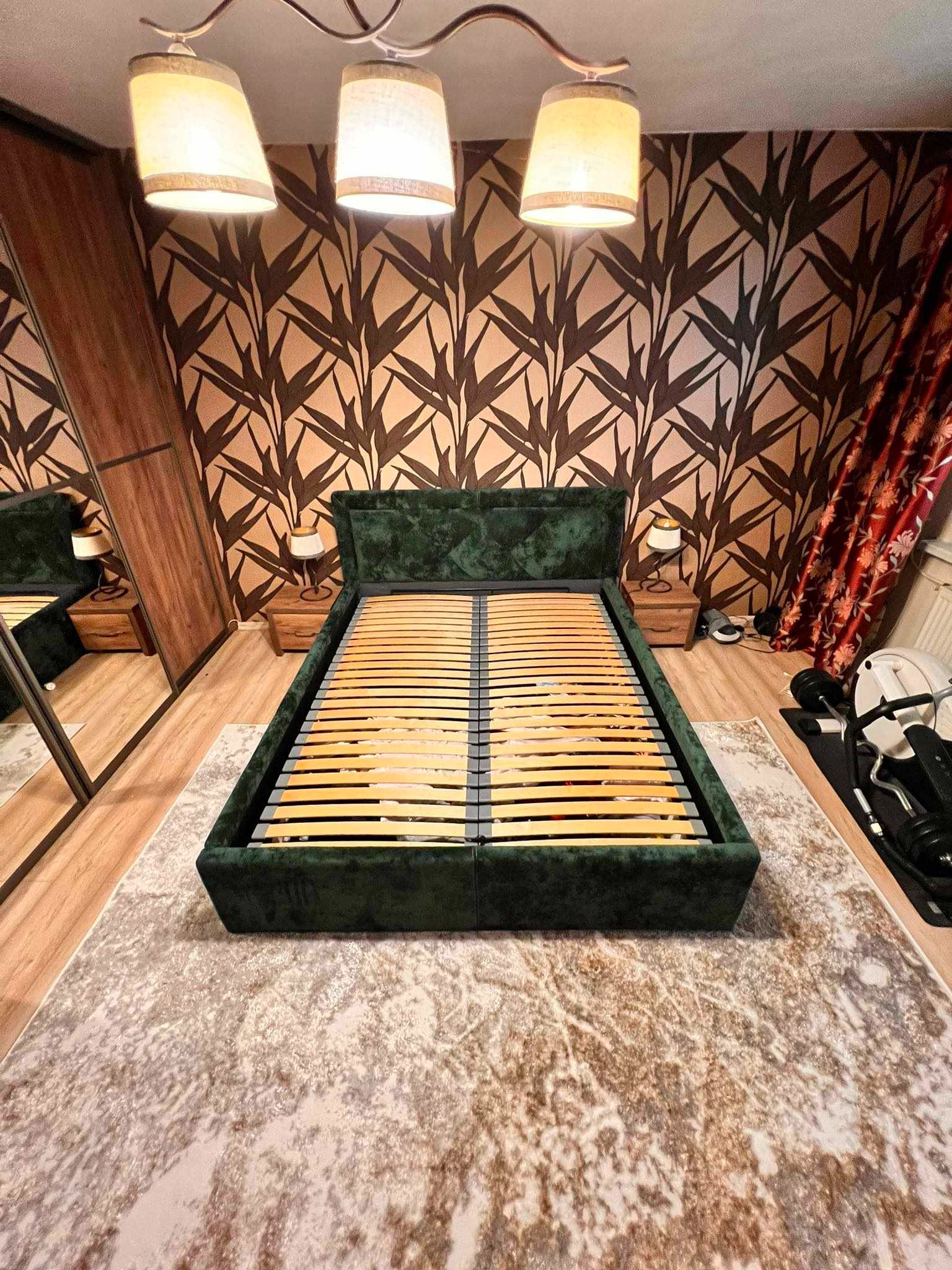 Szmaragdowe łóżko z drewnianym stelażem na tle wzorzystej tapety i zasłon. Widoczne szafki nocne i lampki. Ujęcie z perspektywy podłogi.