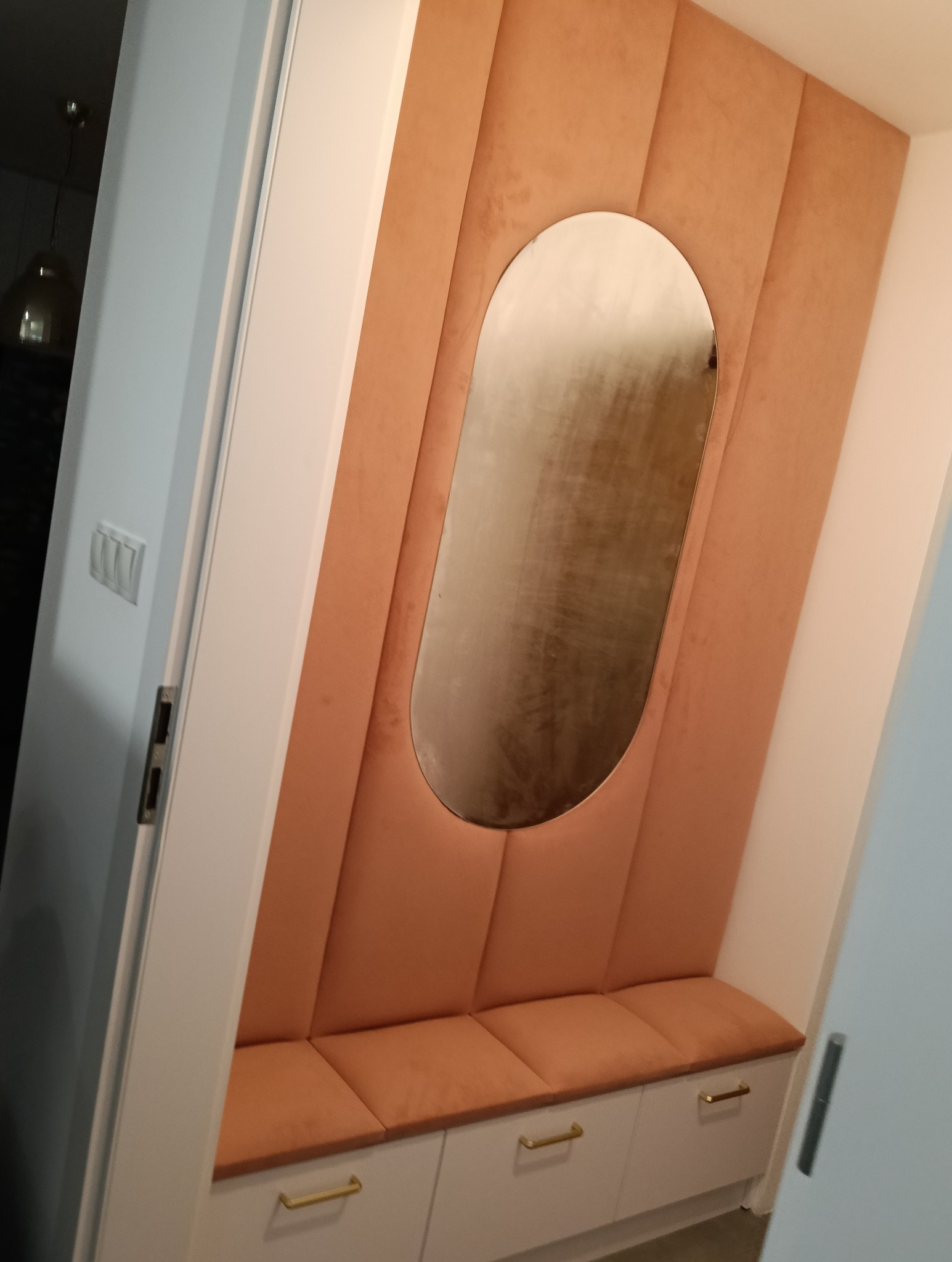 Tapicerowany panel ścienny w odcieniach brzoskwini z owalnym lustrem i siedziskiem z szafkami w przedpokoju. Złote uchwyty dodają elegancji.