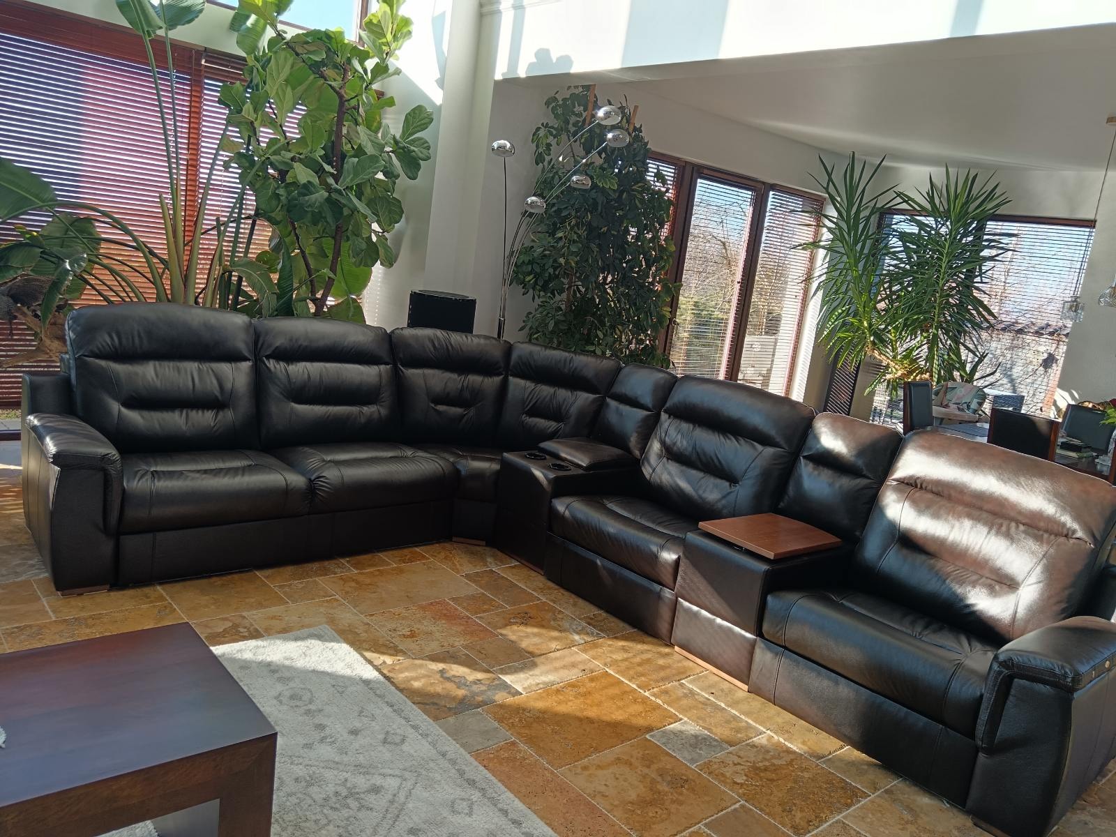 Czarna, skórzana sofa narożna z funkcją relaks w jasnym salonie z dużymi oknami i roślinami. Widoczny stolik kawowy i dywan. Sofa po renowacji.