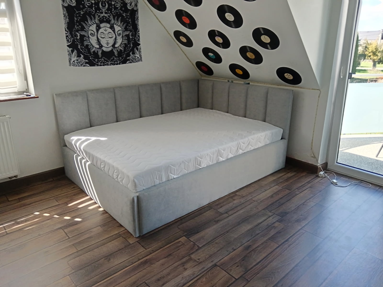 Tapicerowane łóżko w szarym odcieniu, z wezgłowiem w kształcie litery L, dopasowane do skosu dachu. Nad łóżkiem dekoracja ścienna z motywem płyt winylowych i mandali.