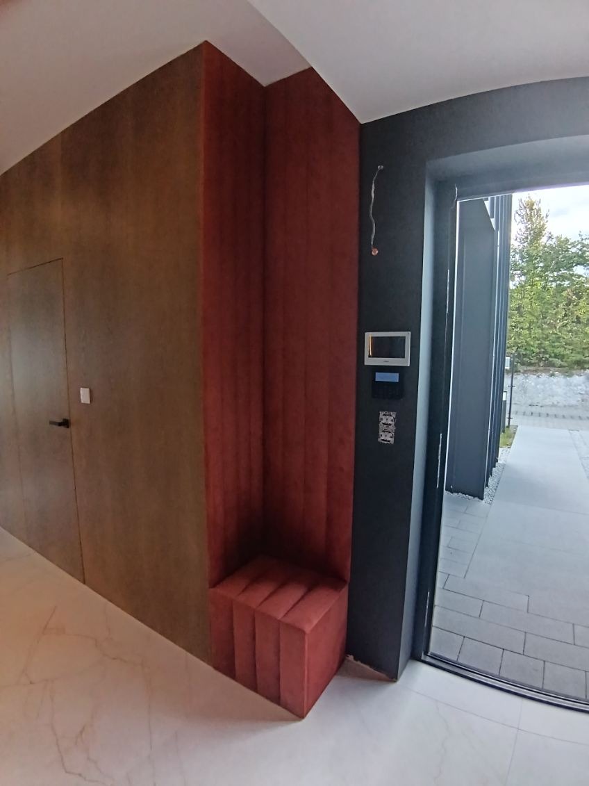 Nowoczesna aranżacja wnętrza: wnęka z siedziskiem obita czerwoną tkaniną, brązowa ściana z ukrytymi drzwiami, marmurowa podłoga i widok na zewnątrz.