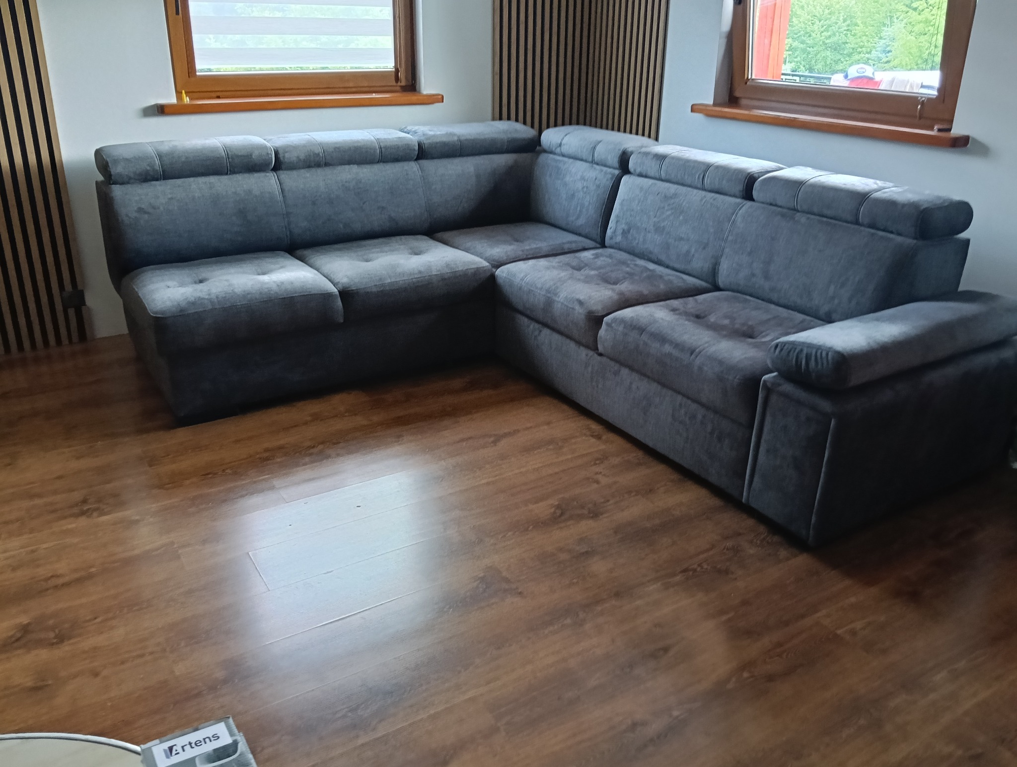 Szara, narożna kanapa z poduszkami na siedzisku, ustawiona w rogu pokoju z drewnianą podłogą i oknami. Widoczne panele dekoracyjne na ścianie.