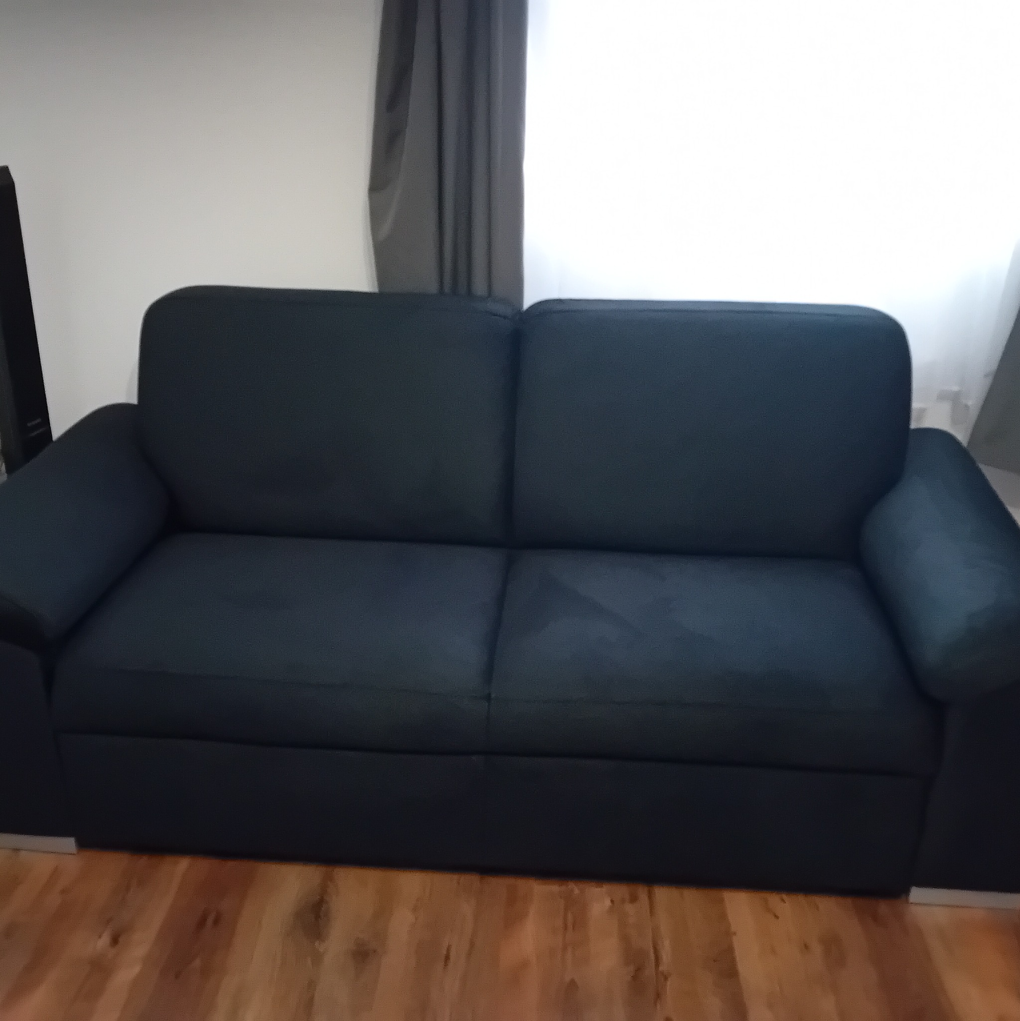 Ciemnogranatowa sofa dwuosobowa z poduszkami na drewnianej podłodze, ustawiona pod białą ścianą z szarą zasłoną w tle. Minimalistyczny design.