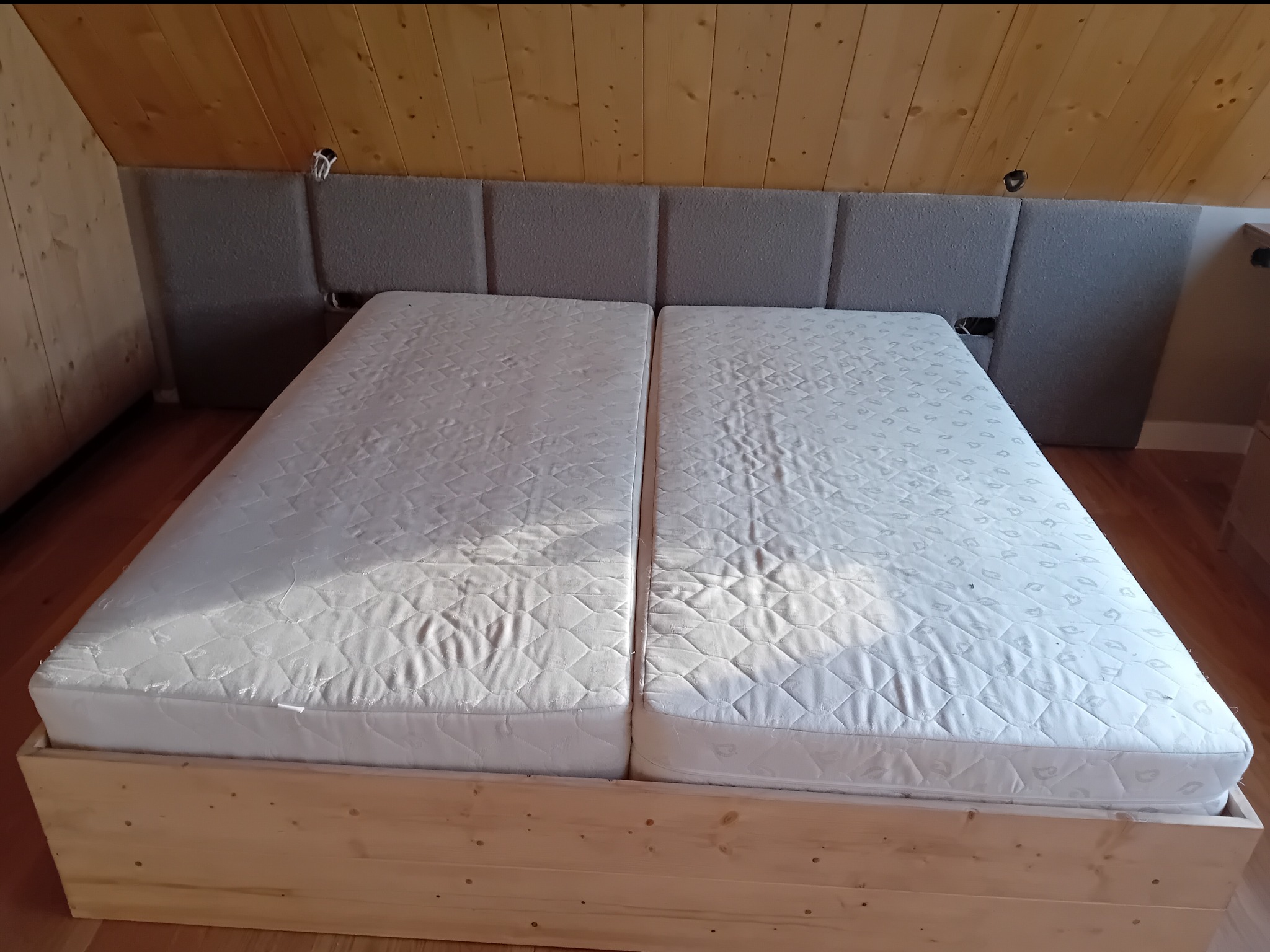 Drewniane łóżko małżeńskie z tapicerowanym wezgłowiem w szarym odcieniu, dopasowane do drewnianej boazerii na poddaszu. Widoczne dwa materace.