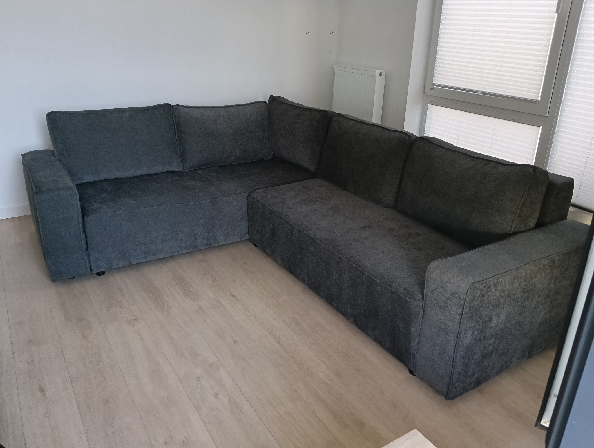 Szara, narożna sofa z poduszkami w minimalistycznym salonie. Widoczne drewniane panele podłogowe i białe ściany. W tle okno z żaluzjami.