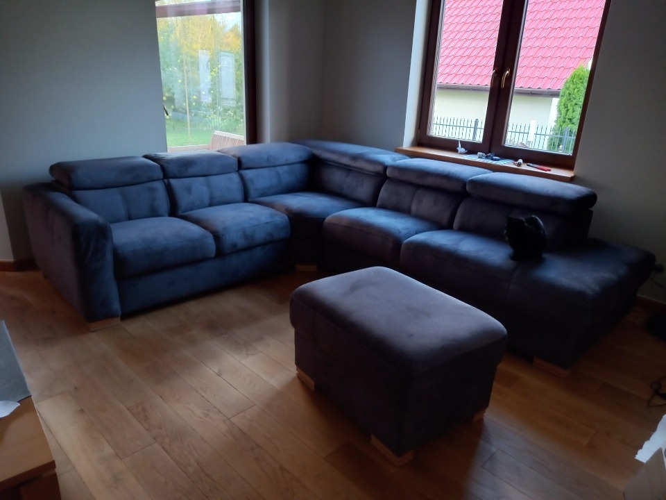 Narożnik tapicerowany w odcieniach szarości z pufą na drewnianej podłodze, z czarnym kotem siedzącym na jednym z segmentów.