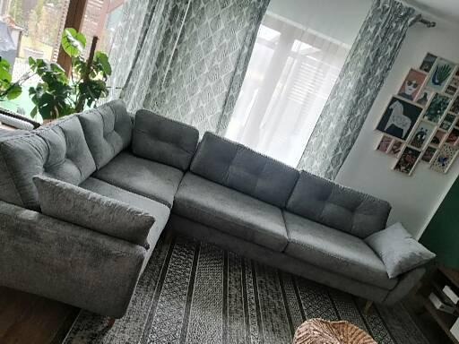 Szary narożnik tapicerowany z guzikami, ustawiony na dywanie w paski, w tle okno z wzorzystymi zasłonami i galeria zdjęć na ścianie.