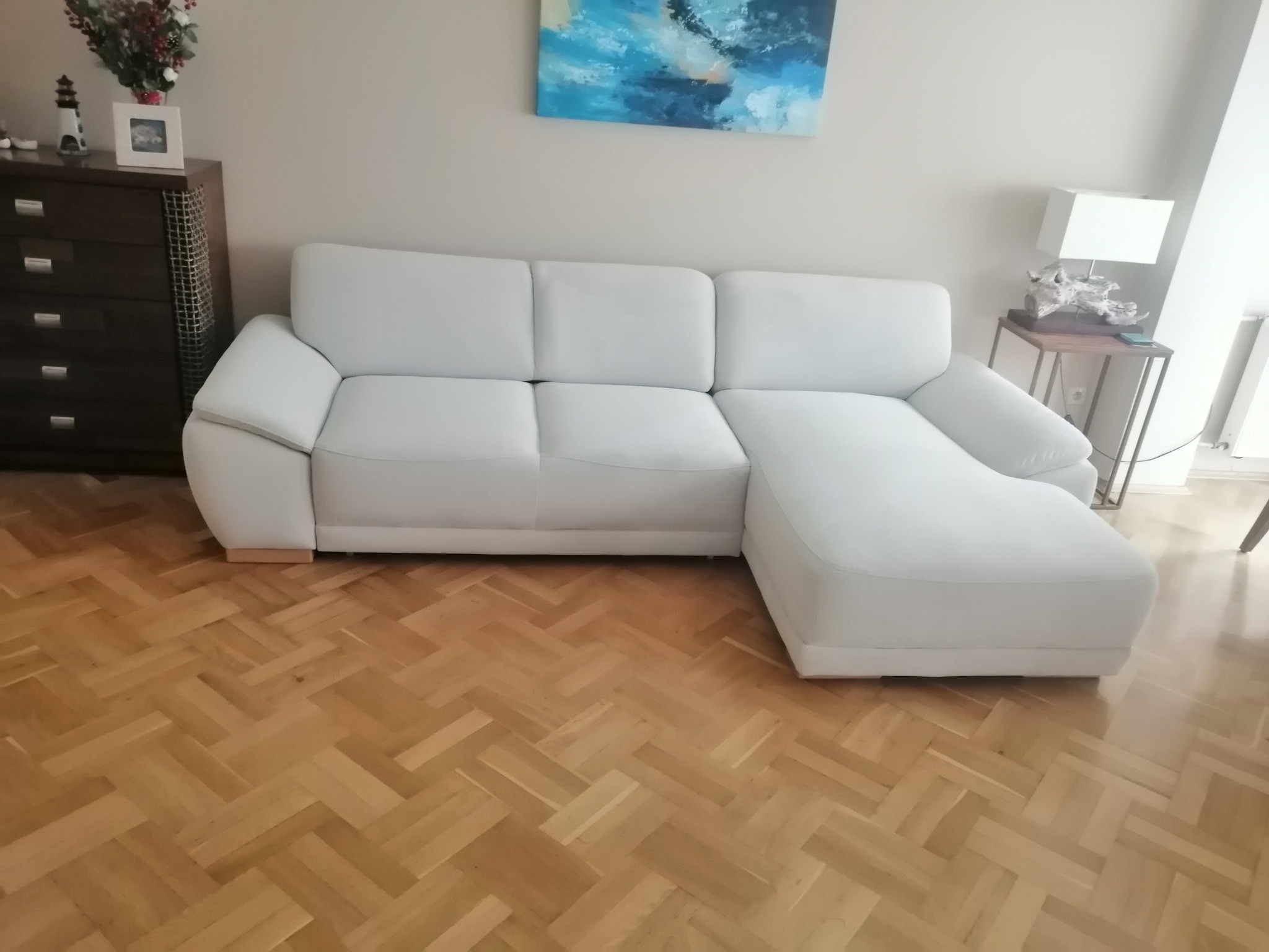 Jasna, skórzana sofa narożna z szezlongiem, ustawiona na parkiecie w jodełkę, z widocznym obrazem abstrakcyjnym na ścianie i komodą z dekoracjami w tle.