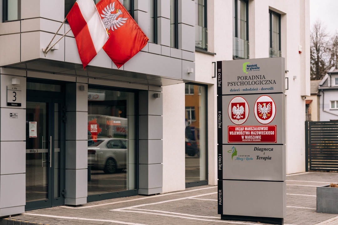 Fasada budynku z szyldem 'Poradnia Psychologiczna' dla dzieci, młodzieży i dorosłych, wiszące flagi Polski, adres budynku Milą 3, widoczny samochód w odbiciu szyby.
