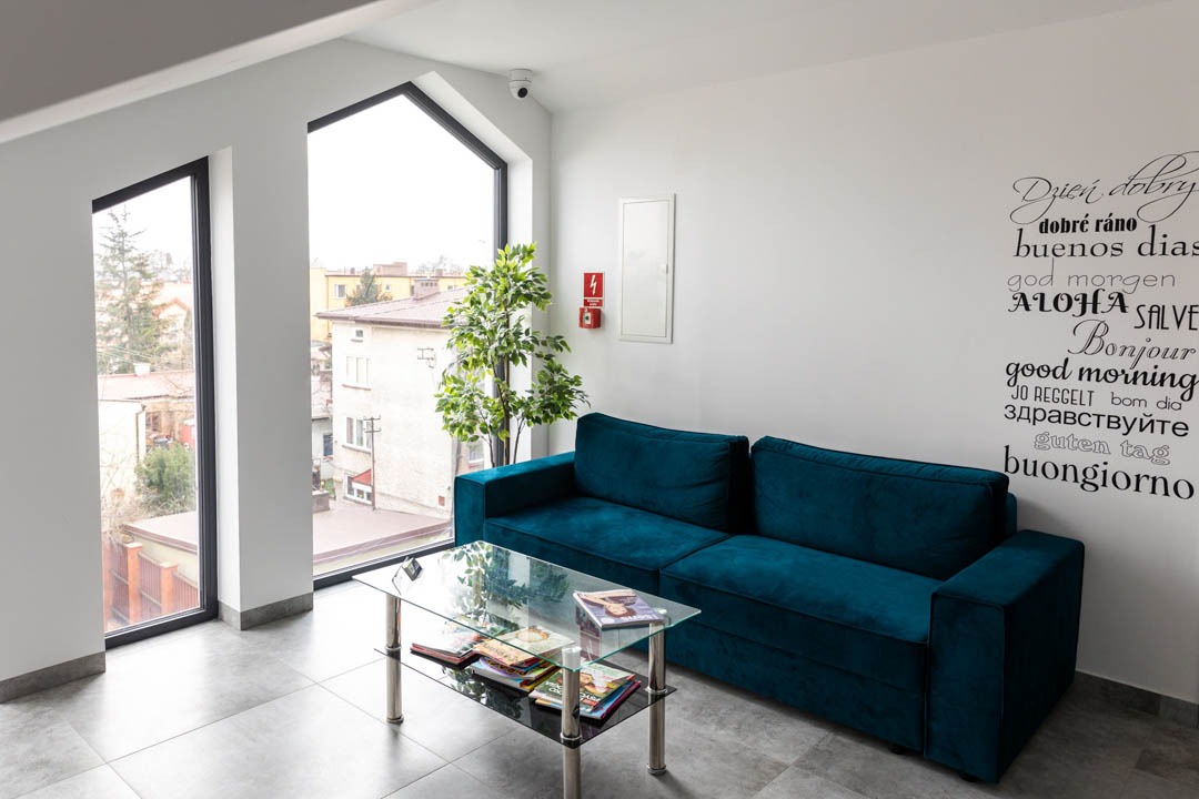Nowoczesne wnętrze poczekalni z turkusową sofą, szklanym stolikiem z czasopismami, dużym oknem z widokiem na miasto i napisem 'Dzień dobry' w różnych językach na ścianie.